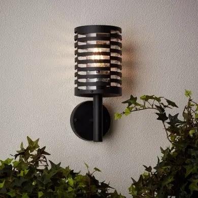 Applique murale extérieure noire avec abat-jour tube perforé et lumière émise vers l’extérieur, fixée au mur près de fleurs